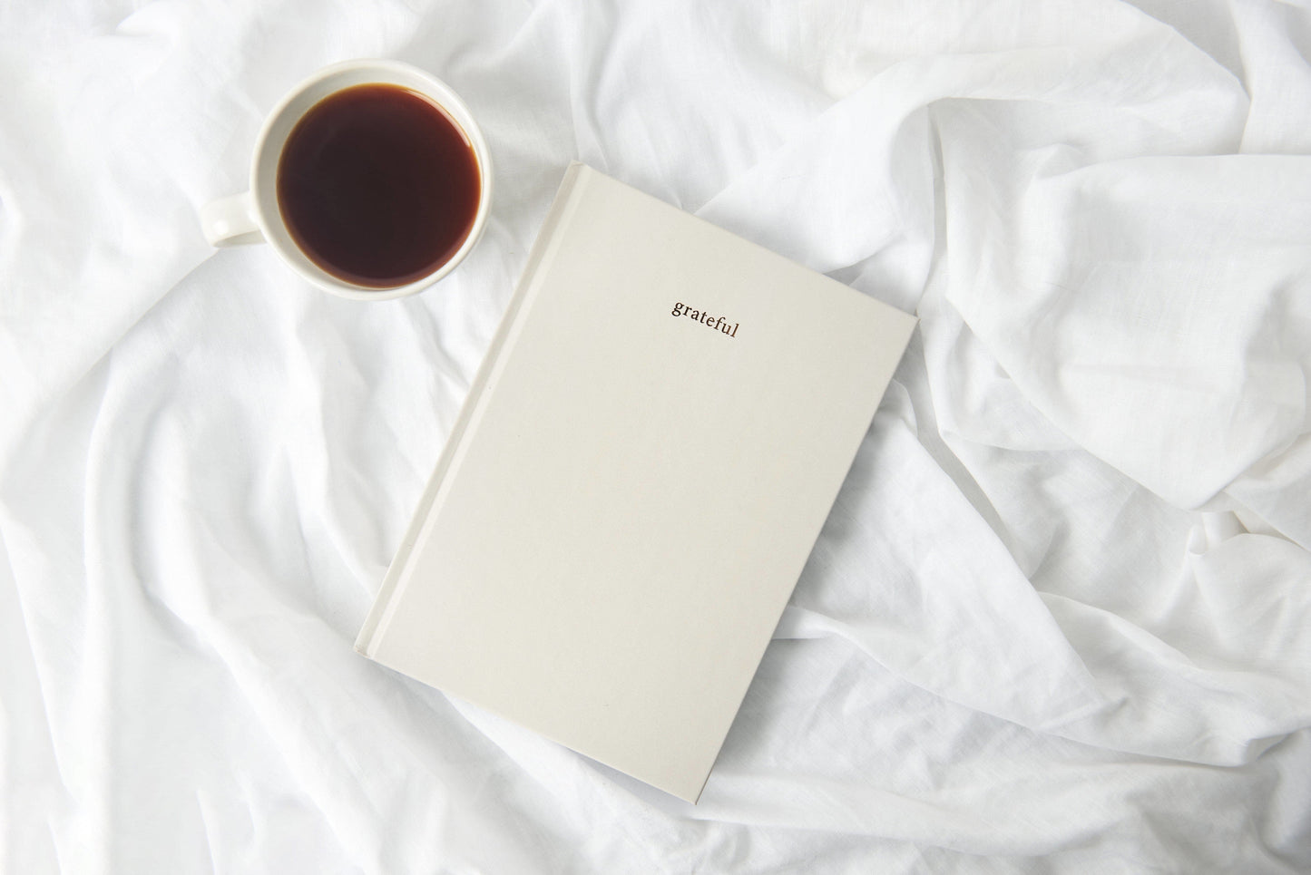A daily gratitude journal sitting on a bed with a cup of coffee beside it